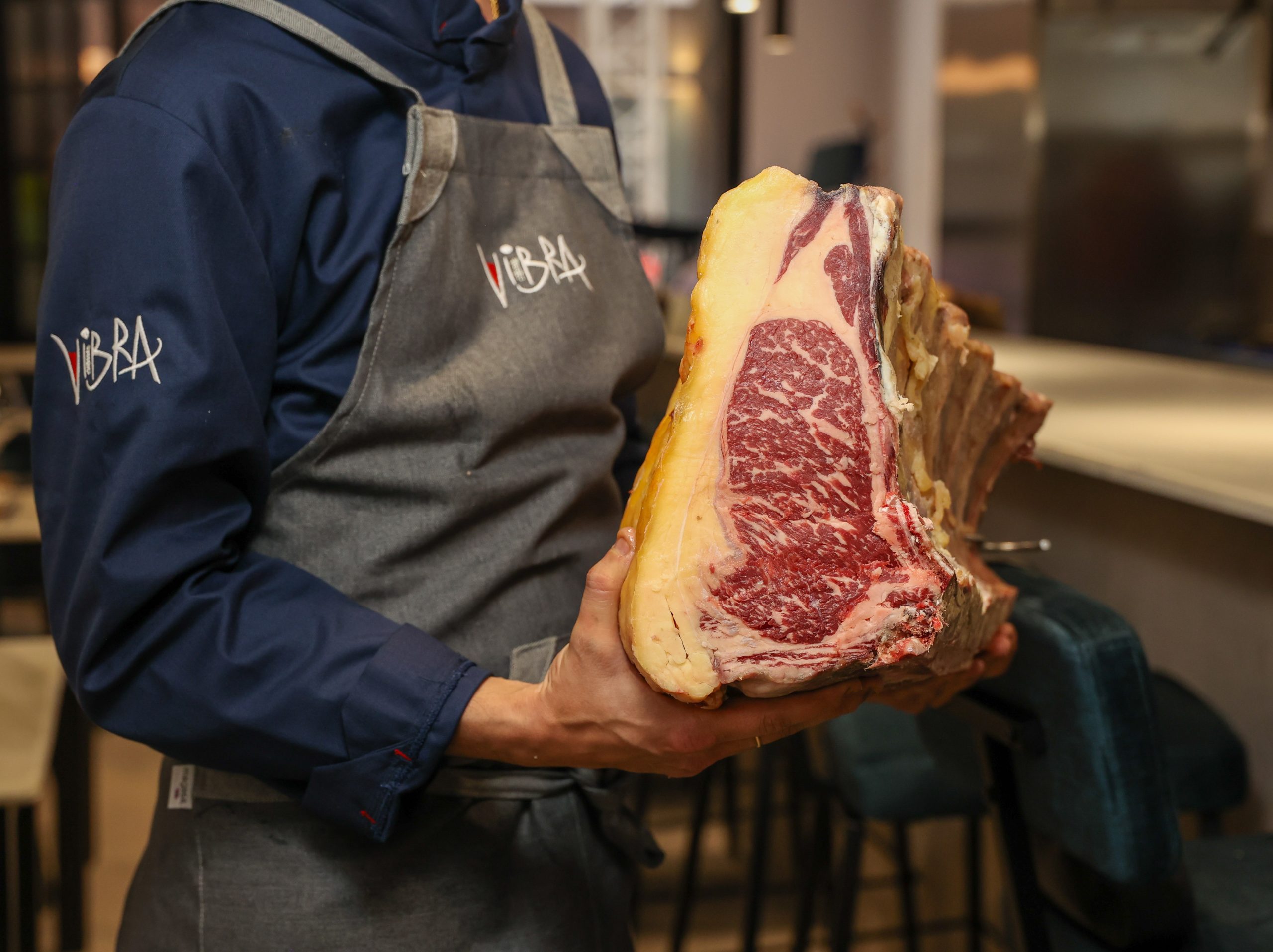 Chef sosteniendo una gran pieza de carne añejada en un restaurante con un delantal de 'Vibra'.