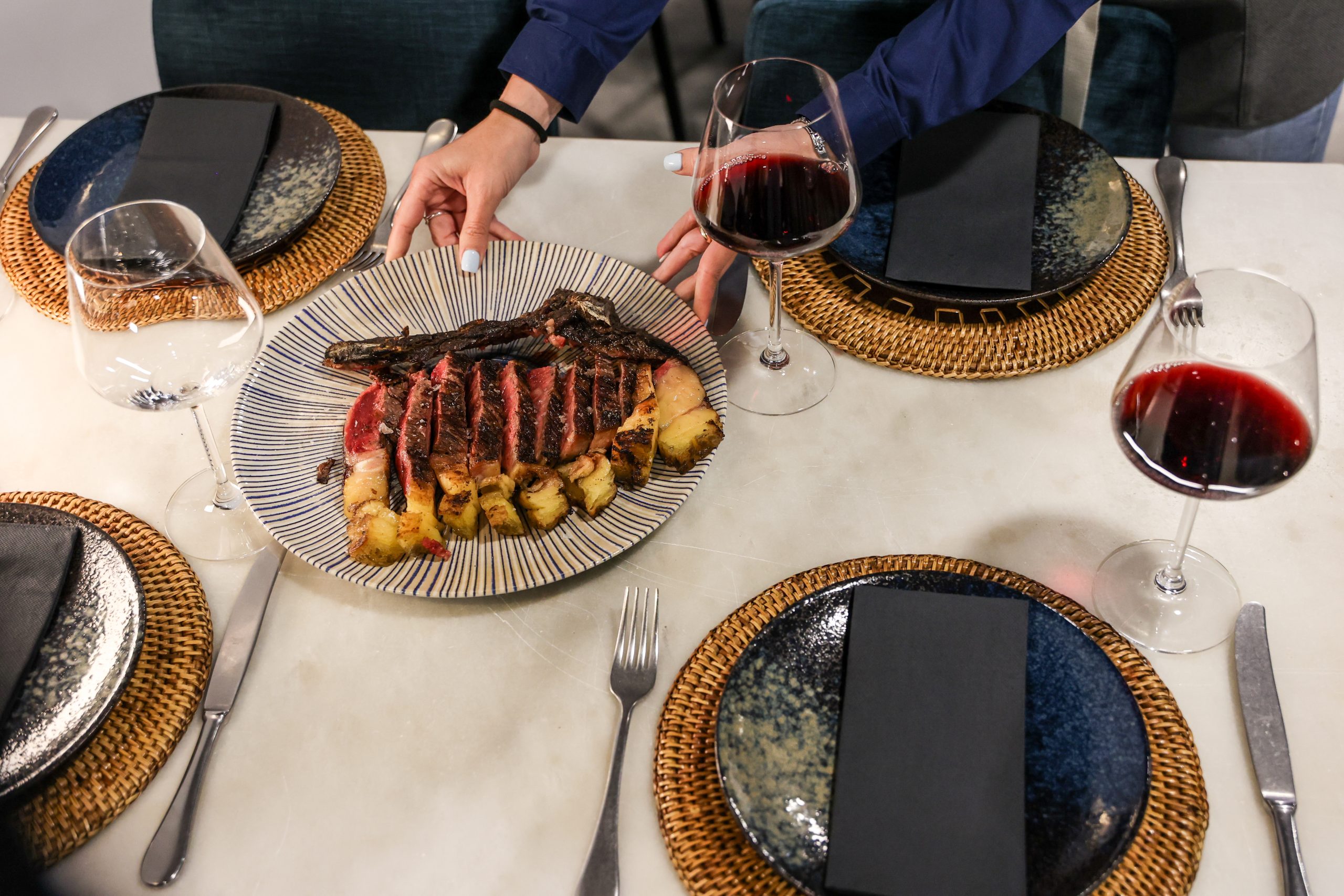 Mano sirviendo un plato de carne a la parrilla en una mesa elegantemente decorada con copas de vino tinto.