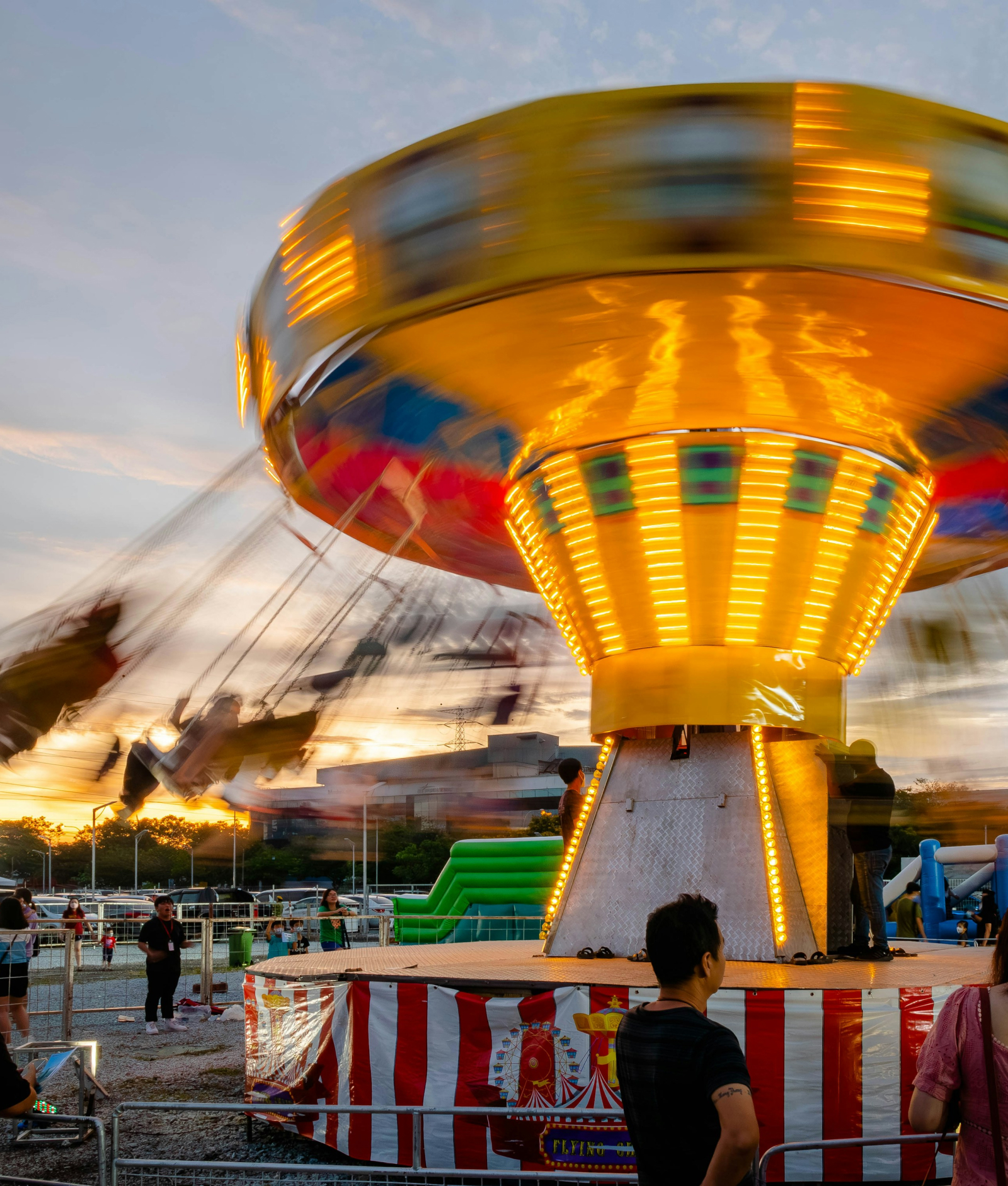 Carrusel giratorio iluminado en movimiento en una feria al atardecer, con luces brillantes y personas disfrutando del ambiente festivo alrededor.